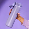A large, light-grey water bottle with a flip-top lid and straw is held by a hand, against a solid purple background.
