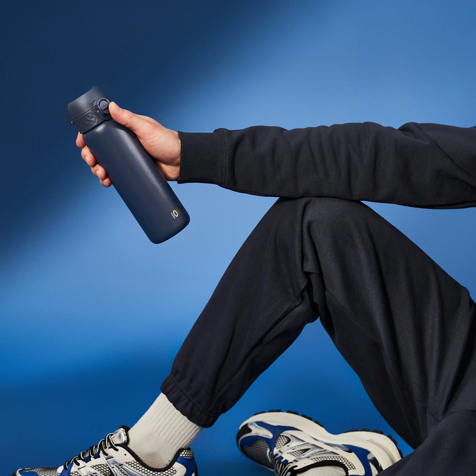 A person in black athletic wear holds a dark blue water bottle in a seated position against a solid blue background wearing white socks and sneakers.