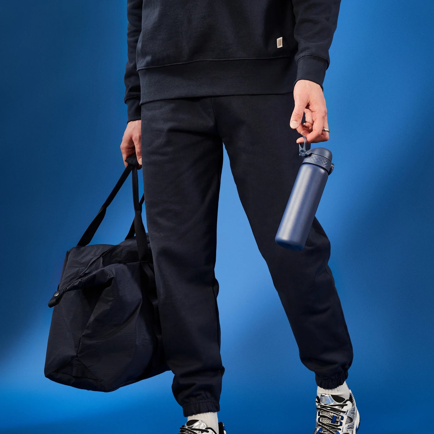 Person holding a dark bag and a blue water bottle against a blue background wearing black sweatpants and sneakers