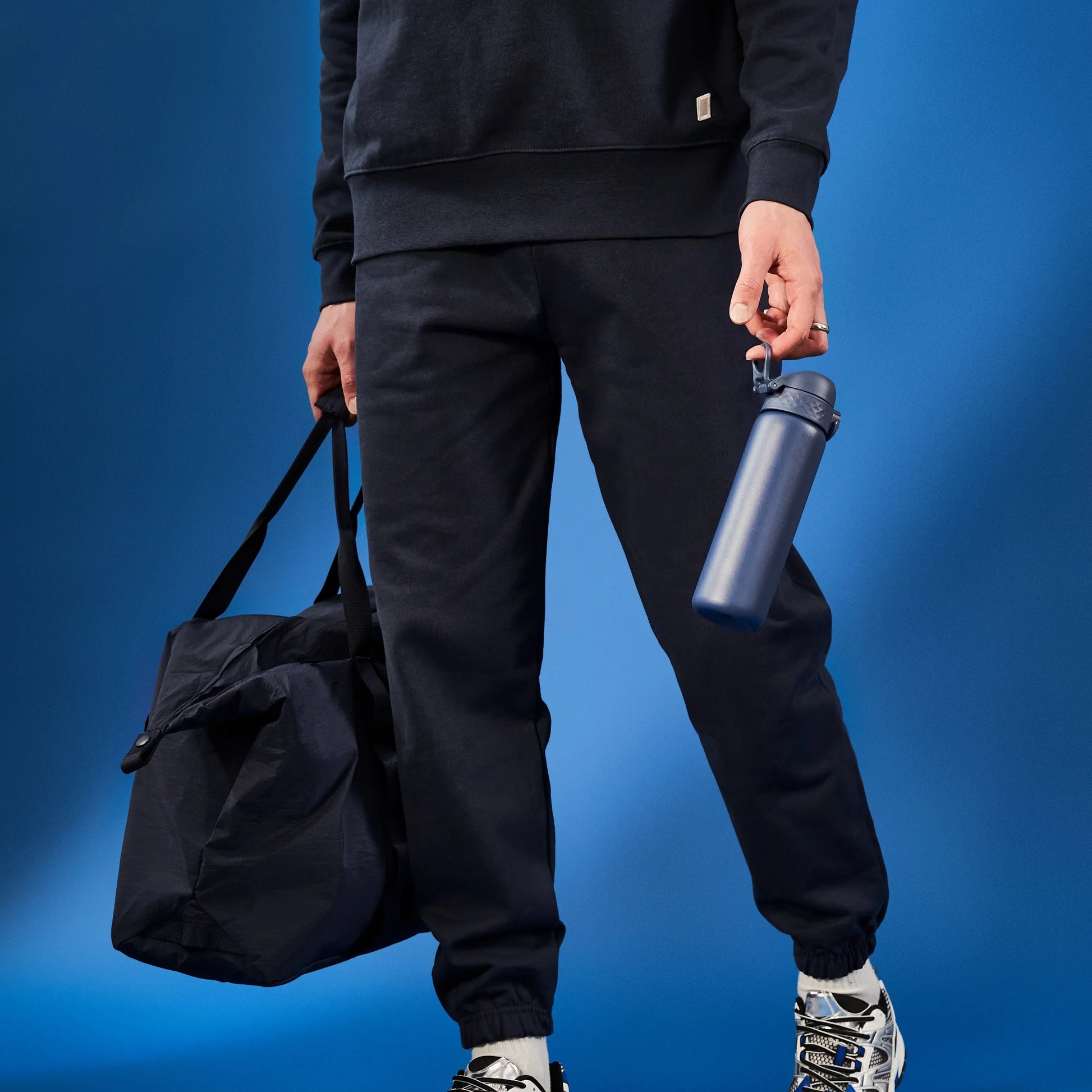 Person holding a dark bag and a blue water bottle against a blue background wearing black sweatpants and sneakers