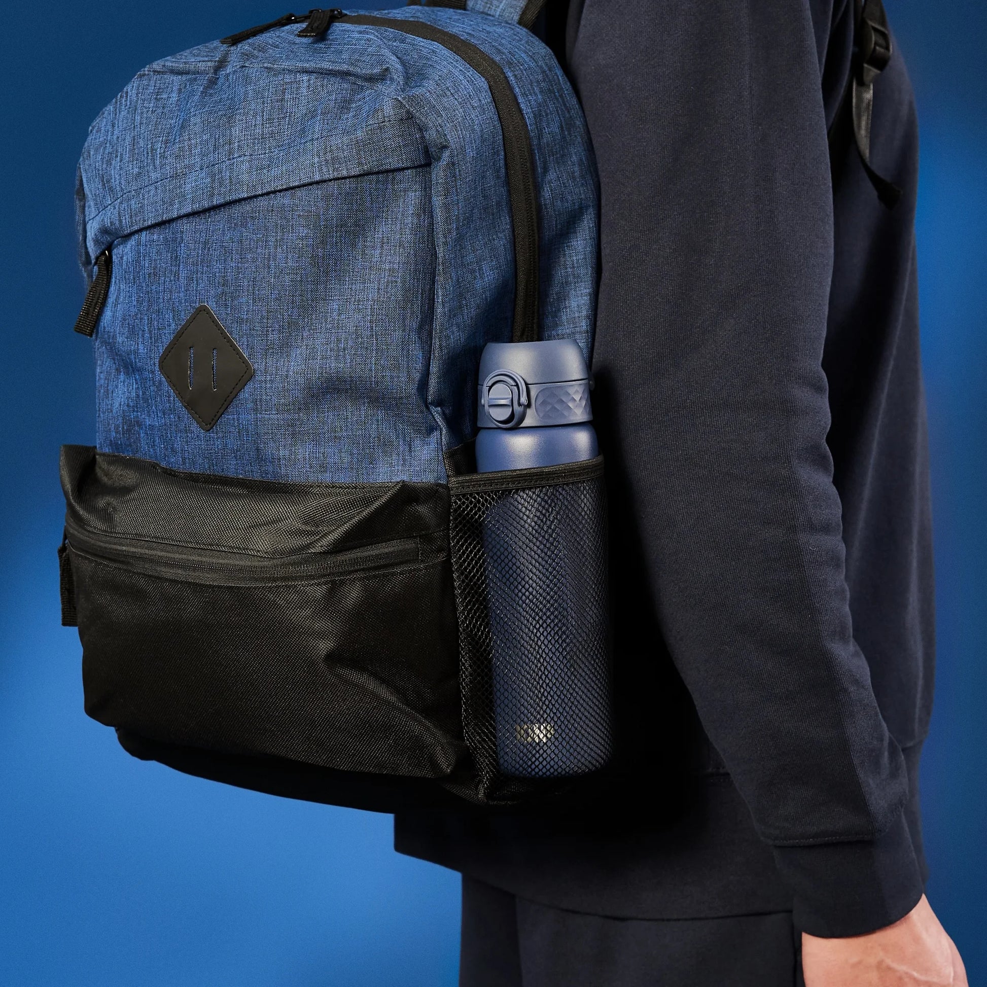 A blue backpack with a black pocket holds a blue water bottle in a mesh side pouch worn by a person dressed in dark clothing against a blue background.