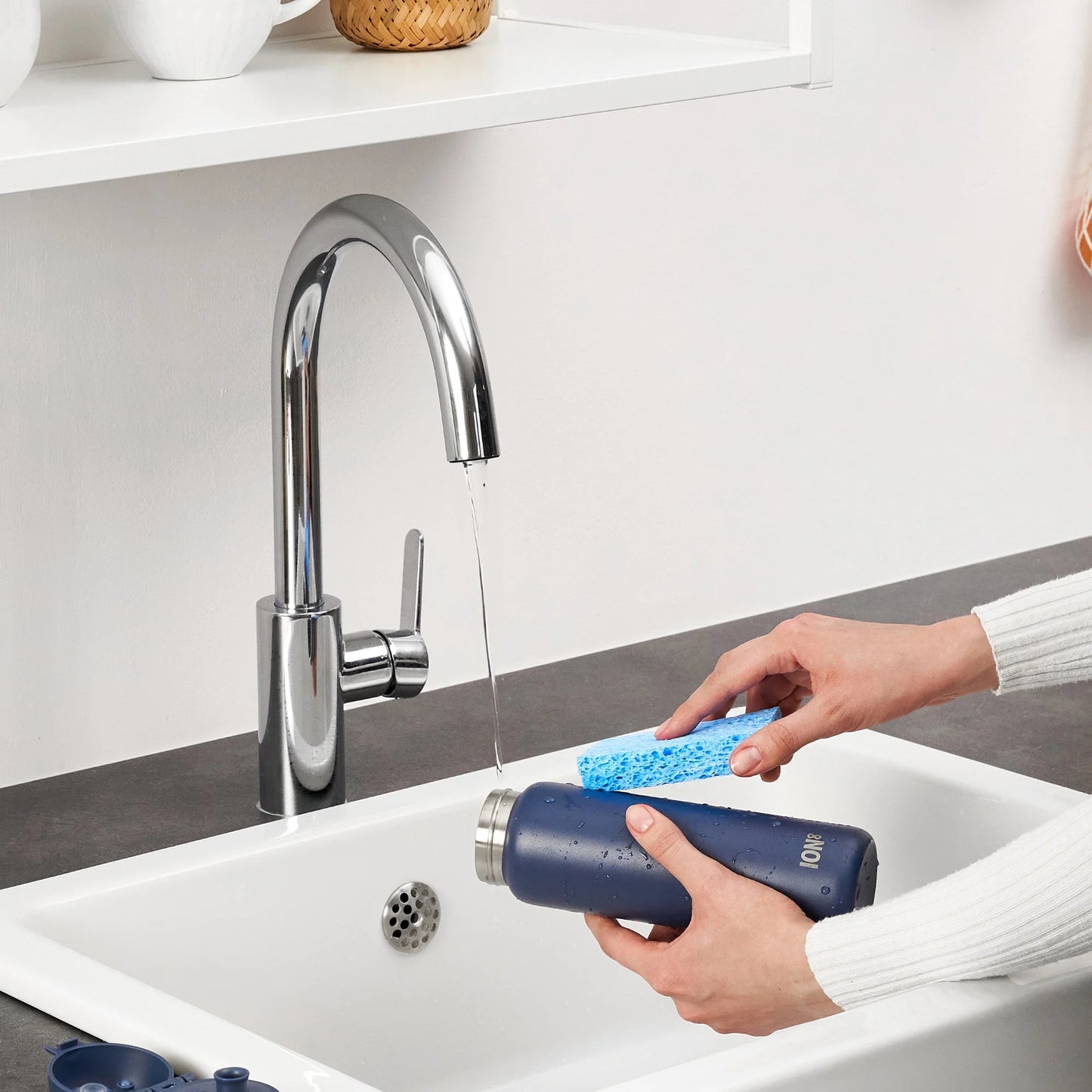 Hands scrubbing a navy bottle with a blue sponge under a running faucet in a kitchen sink context.