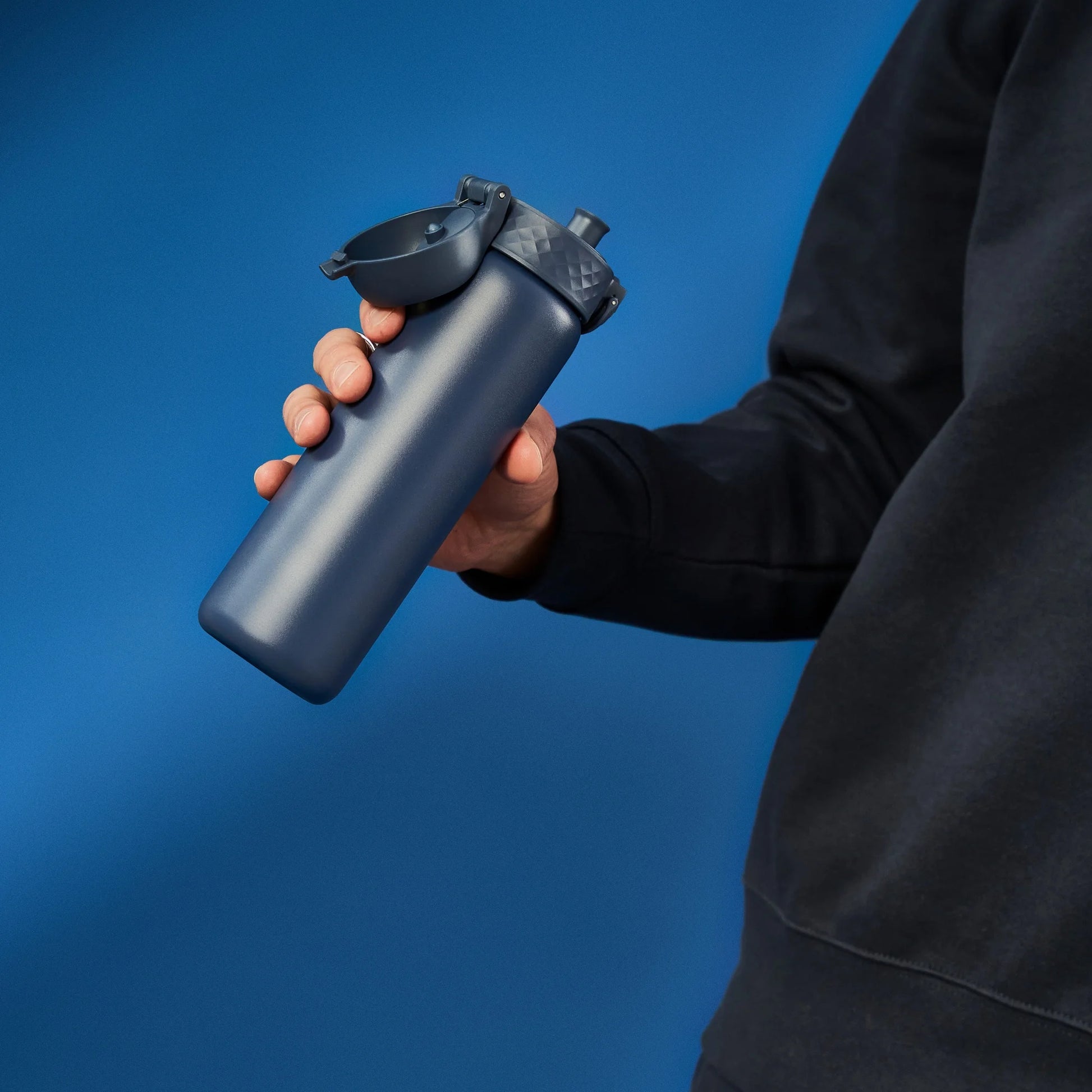 A person holds a blue water bottle with a flip-top lid against a solid blue background wearing a dark sweatshirt.