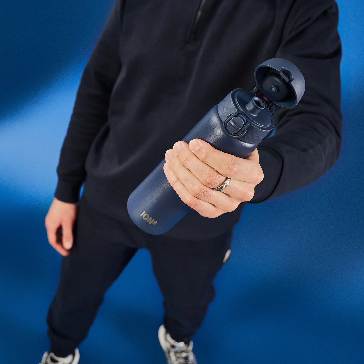 A person holds a dark blue water bottle with a flipped-open lid labeled "ION8" against a blue background wearing a dark outfit.