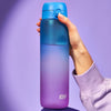A blue and purple ombre water bottle is being held against a light purple background. The bottle has a blue lid with a button.