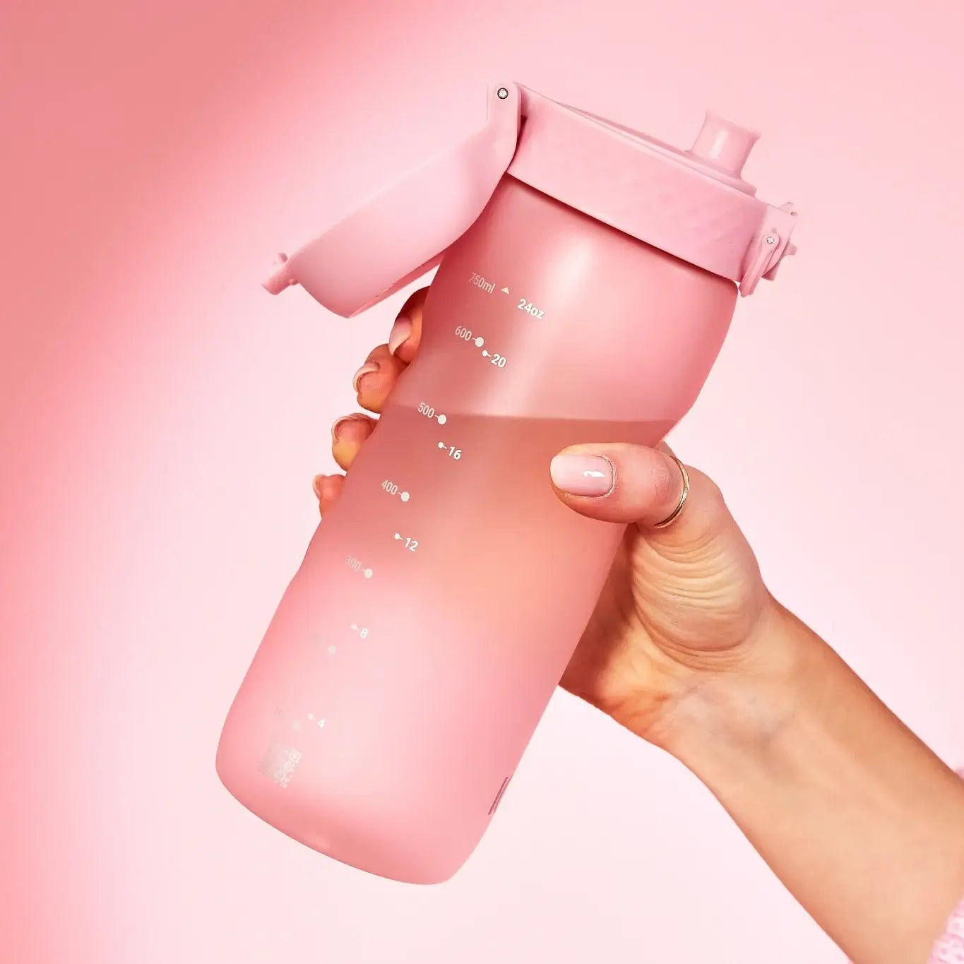 A pink water bottle with a flip-top lid is being held; measurements in milliliters and ounces are visible on its side. The background is a matching pink. 750ml 24oz 600 20 500 16 400 12
