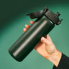 A dark-green, cylindrical water bottle is being held, its flip-top lid slightly ajar, against a muted green background.