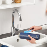 A dark-blue water bottle is being washed with a blue sponge under a chrome faucet in a white kitchen sink. The bottle is labeled 