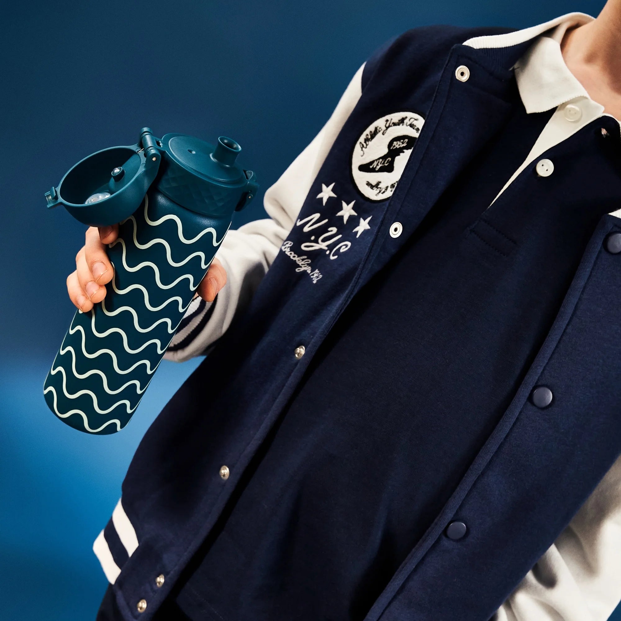 A person holds a water bottle with a wavy pattern open, wearing a blue and white varsity jacket with text 