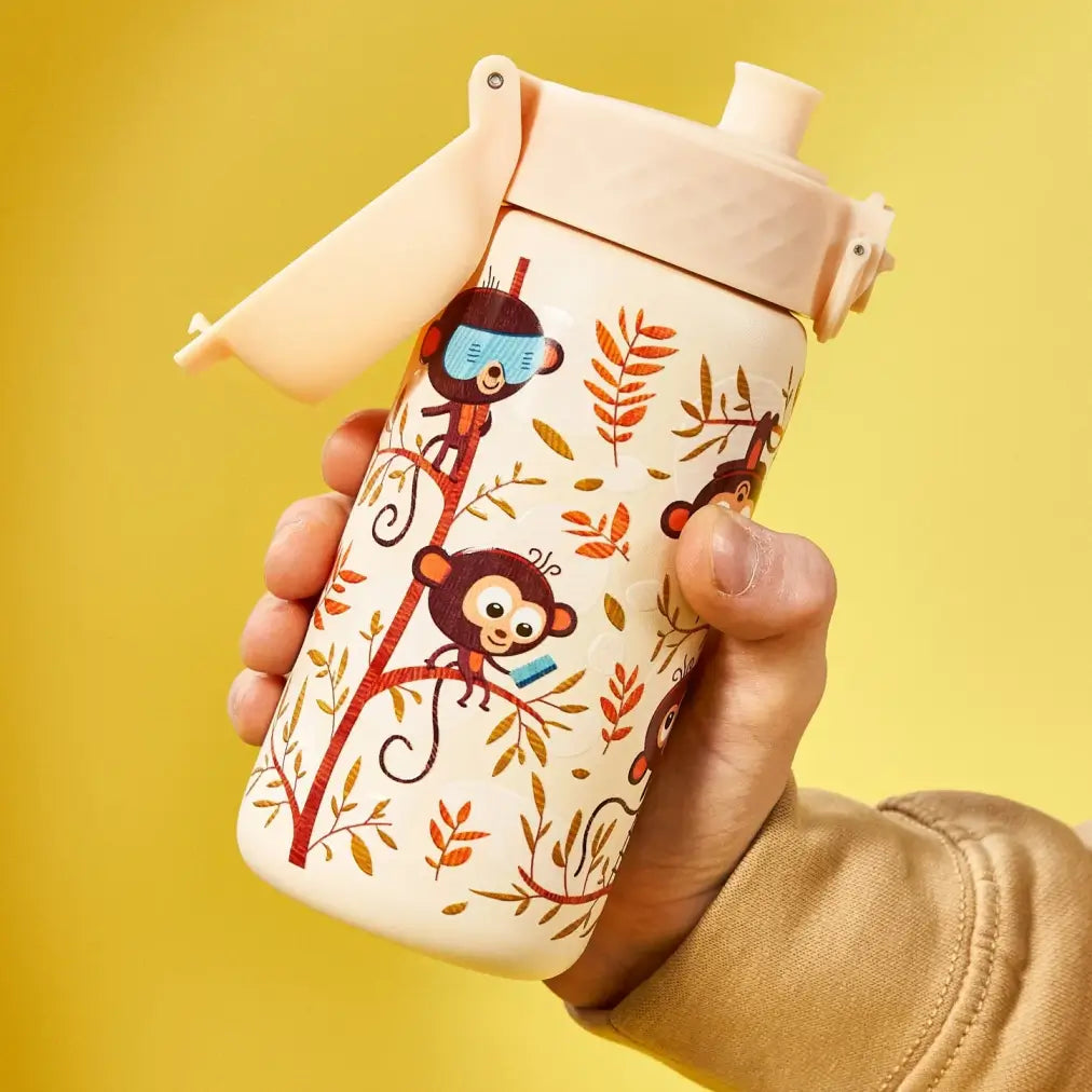 A child's water bottle, featuring a flip-top lid and playful monkey illustrations, is held in a hand against a yellow background.
