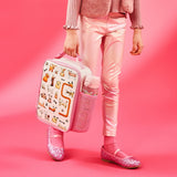 Person carrying a pink lunchbox decorated with cartoon dogs walking in a pink outfit including shiny leggings and glittery shoes against a pink background
