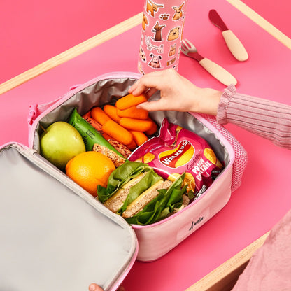 Lunchbox filled with green apple, orange, sandwich, granola bars, carrots, and Walkers crisps. A hand reaches for carrots. Nearby is a decorated water bottle on a pink table. The name "Amelia" is on the lunchbox.