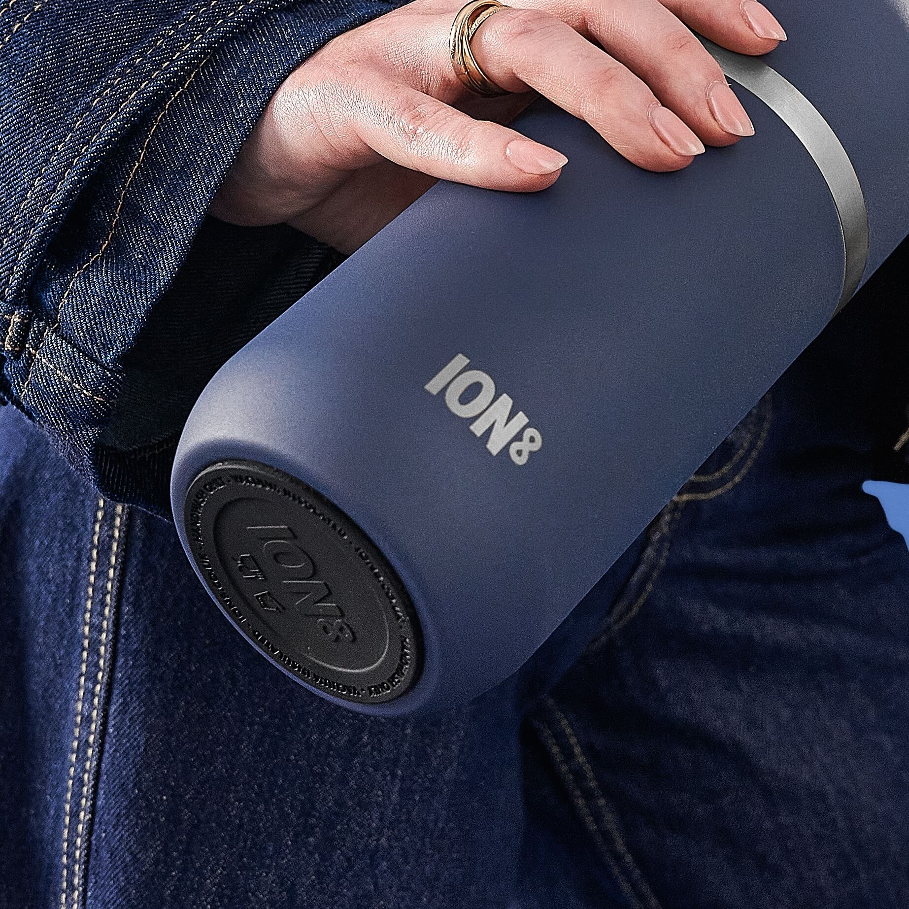 A dark-blue ION8 water bottle is held by a hand, which is wearing rings. The hand and bottle rest against dark denim fabric.