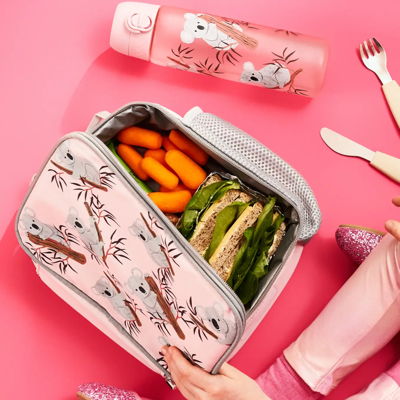 A pink koala-patterned lunch bag containing carrots and a sandwich is being held open; it rests on a pink surface with a matching water bottle and cutlery nearby.