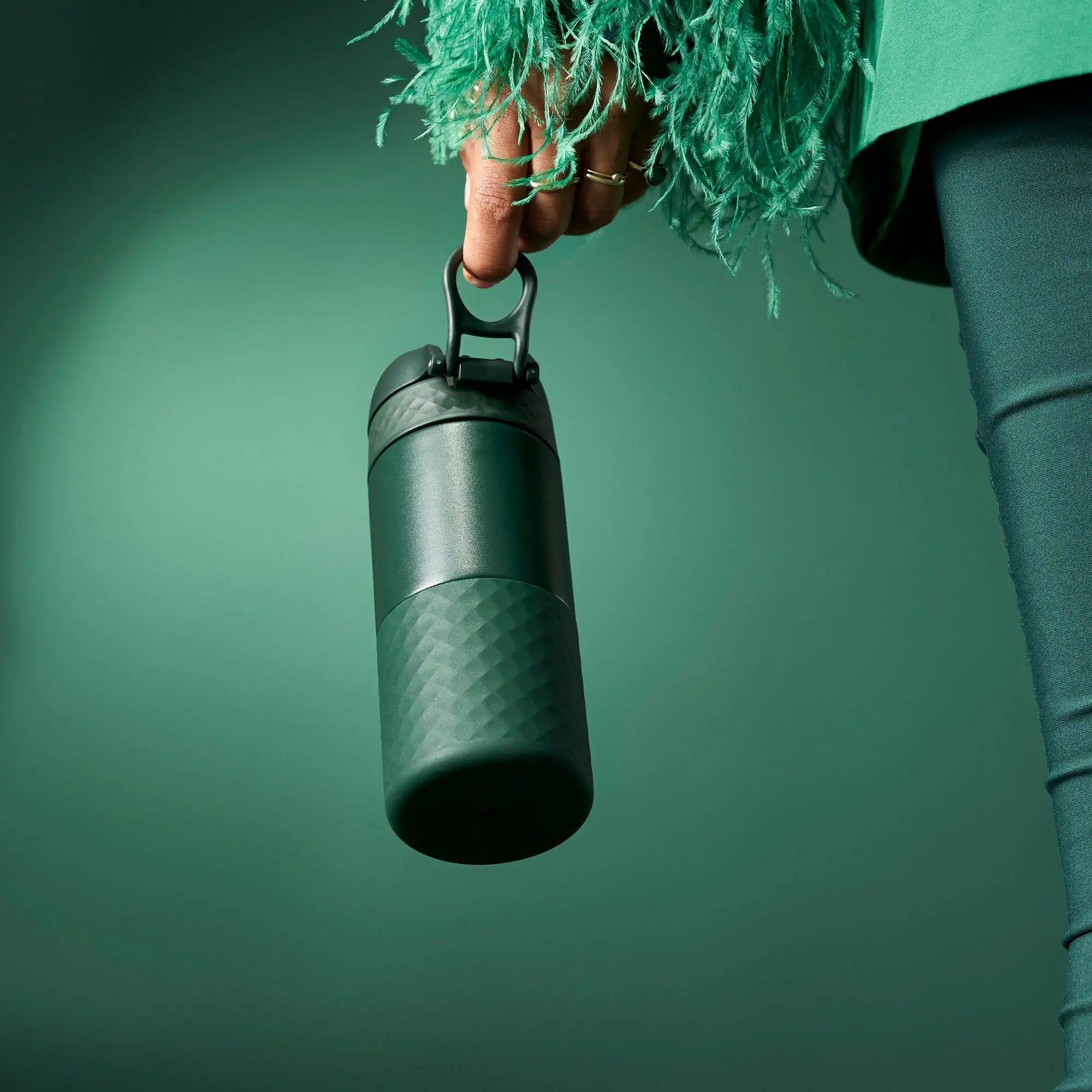 A green insulated bottle is held by a hand wearing a garment with green feathers against a green background creating a monochromatic aesthetic.