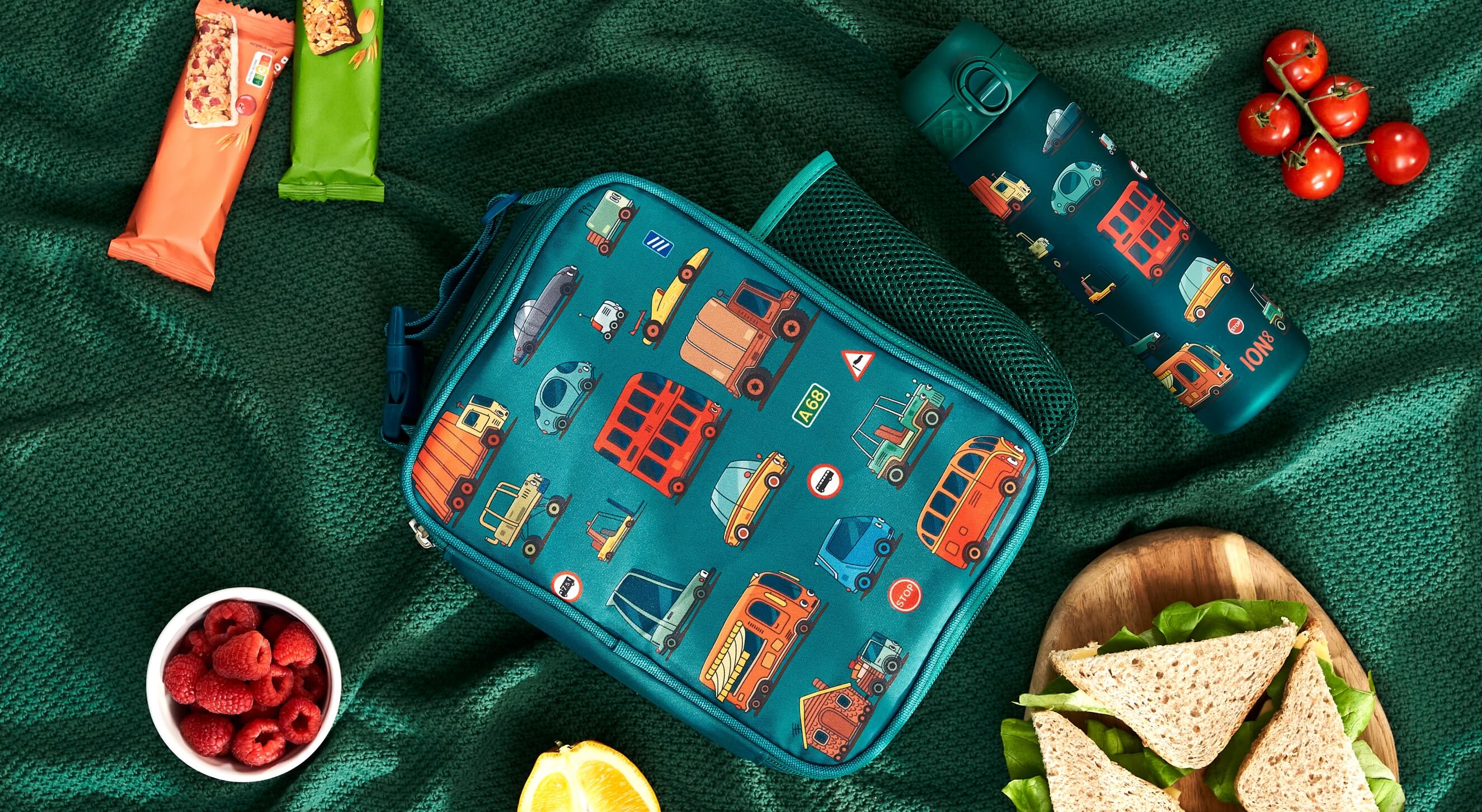 A teal lunch bag, adorned with vehicle illustrations, rests on a green fabric. Nearby are food items and a matching water bottle labeled "ION8".