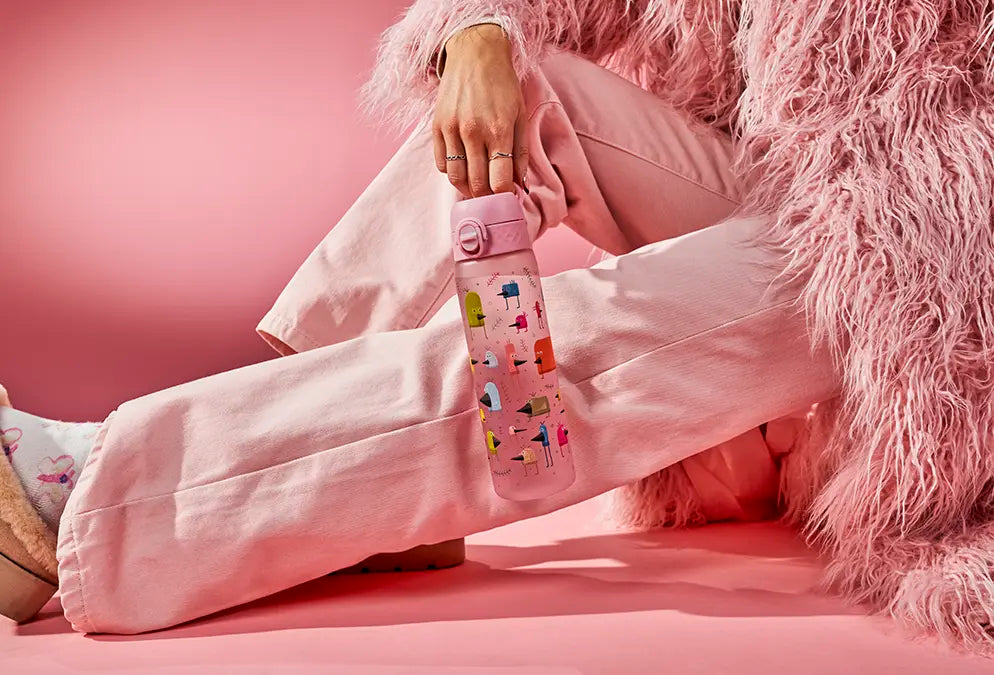 A pink water bottle, adorned with cartoon birds, is held by a person wearing pink pants and a fluffy pink coat, sitting on a pink surface.