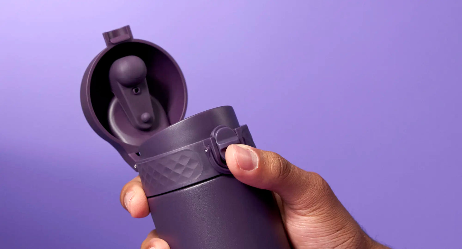 A purple travel mug is being held and opened against a purple background. A person’s hand is shown interacting with the lid’s mechanism.