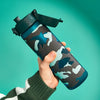 A camouflage-patterned water bottle is being held; a person’s hand grips it against a teal backdrop.