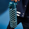 A teal water bottle, featuring a wavy pattern, is being held against a dark-blue backdrop. The bottle's brand name, "ALO," is subtly visible on its side.